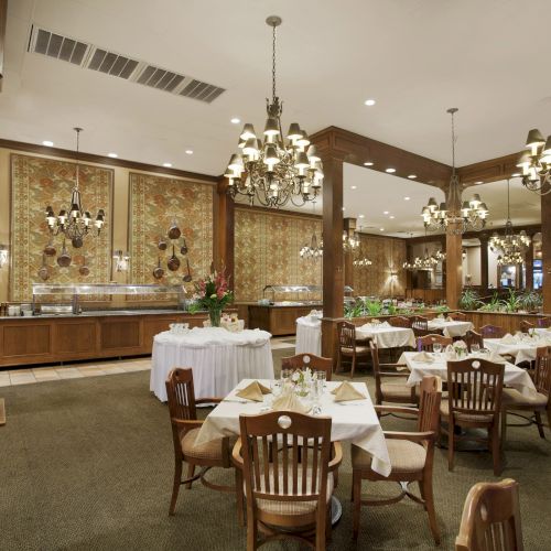 A spacious, elegant dining room with set tables, chandeliers, and a buffet area, featuring wooden decor and upholstered chairs.