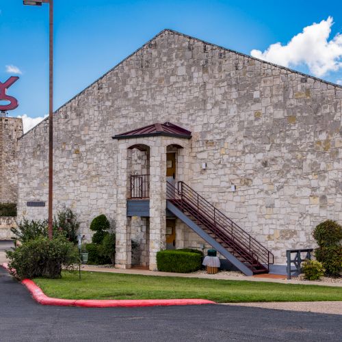 The image shows a stone building with red-accented outdoor stairs, a green lawn, and a sculpture in the background under a blue sky.
