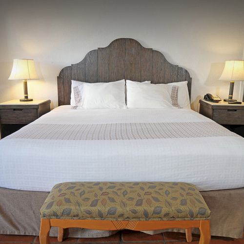 The image shows a neatly made bed with a decorative headboard, two lamps on bedside tables, and a bench at the foot.