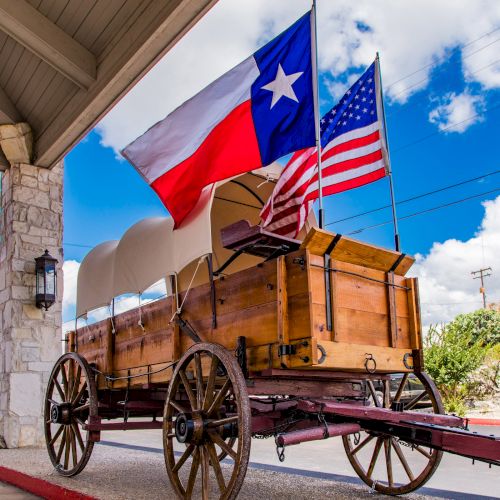 A wooden wagon on display with Texas and American flags, set under a porch against a bright blue sky with clouds.