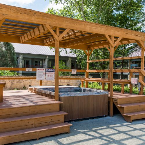 A wooden pergola covers a hot tub with stairs and railings, surrounded by greenery and buildings in the background.