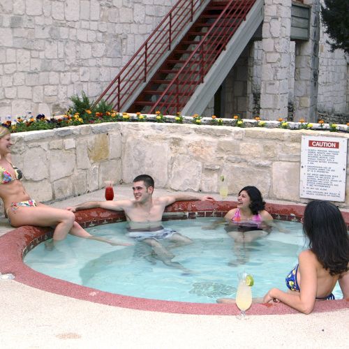 Four people relaxing in a hot tub, each holding drinks, with a stone wall and staircase in the background.