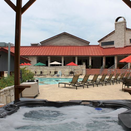 A hot tub in the foreground, overlooking a pool area with lounge chairs and red umbrellas, set against a building with a red roof.
