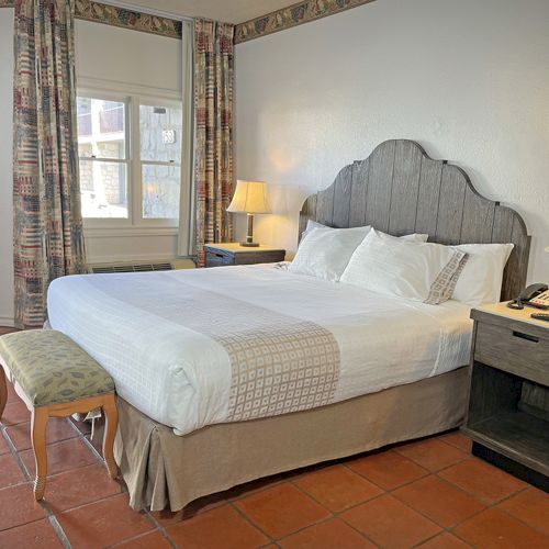 The image shows a cozy hotel room with a large bed, two side tables with lamps, a window, and decorative elements on the walls.