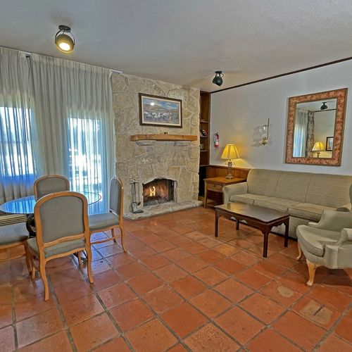 A cozy living room with a stone fireplace, sofa, armchair, dining table, art on the walls, and terracotta tile flooring.
