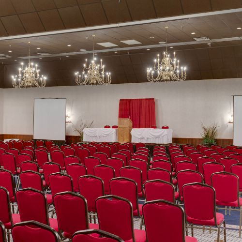 A conference room with red chairs, two projection screens, chandeliers, and a podium with a red backdrop is shown in the image.