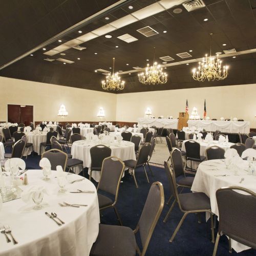 A banquet hall is set up with round tables, white tablecloths, and chairs; chandeliers hang from the ceiling, creating an elegant atmosphere.