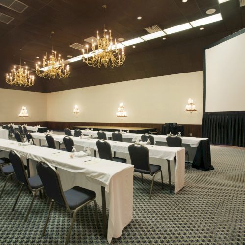 A conference room with rows of tables and chairs, chandeliers, and a large screen for presentations is shown.