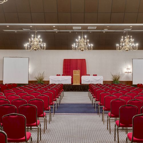 An empty conference room with red chairs, chandeliers, two projector screens, and a podium at the front.
