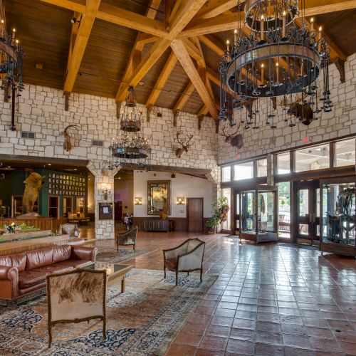 This image shows a spacious rustic lobby with wooden beams, chandeliers, stone walls, leather seating, and large windows.