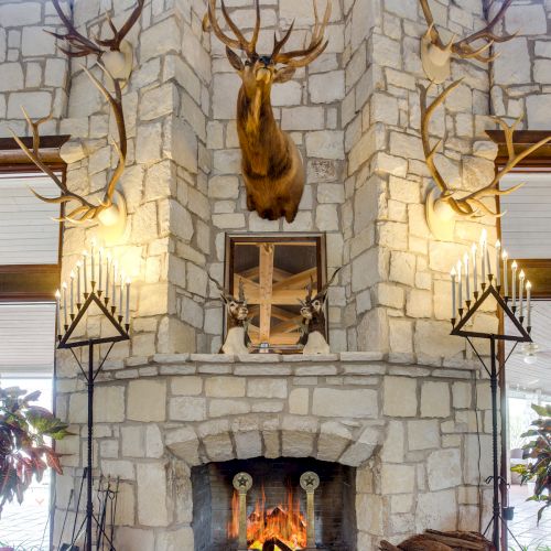A stone fireplace with mounted deer antlers, a roaring fire, candles, and decorative logs on a stone hearth.