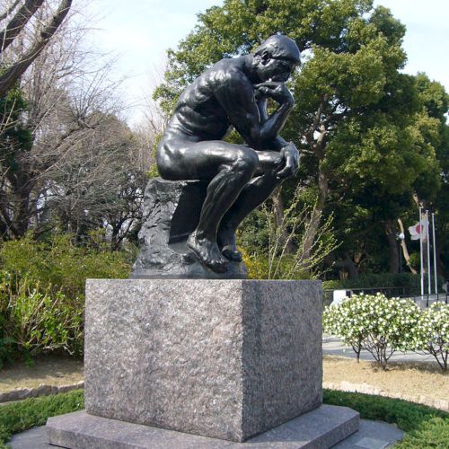 The image shows a statue of a man sitting and thinking, posed with his chin resting on one hand, surrounded by a park landscape.