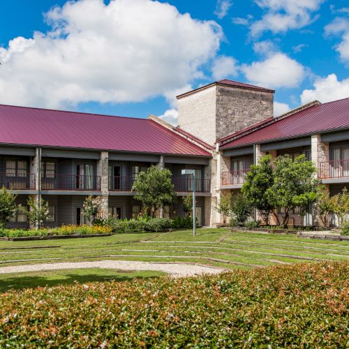 A two-story building with a red roof and stone exterior is surrounded by trees, shrubs, and a manicured lawn under a blue sky with clouds.
