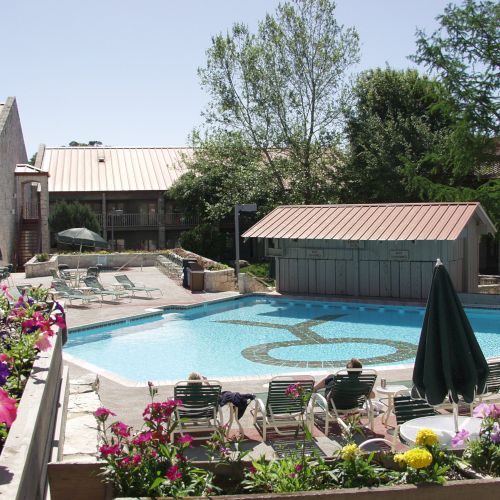 The image shows a swimming pool area surrounded by buildings, flowers, and greenery, with pool chairs and umbrellas arranged around it.