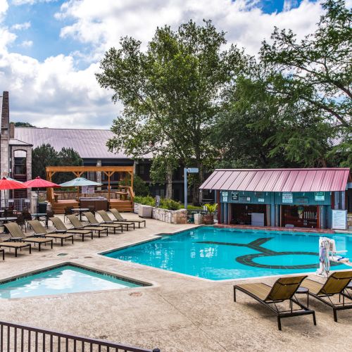 A swimming pool area with loungers, red umbrellas, and a small building, surrounded by trees and structures under a partly cloudy sky.