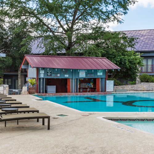 The image shows a swimming pool area with lounge chairs, a poolside bar or building, and surrounding trees and structures.