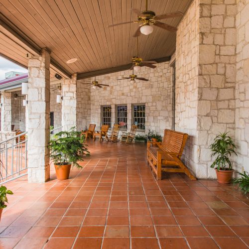 A covered patio with stone walls, tiled floor, wooden seats, ceiling fans, and potted plants placed along the sides of the seating area.