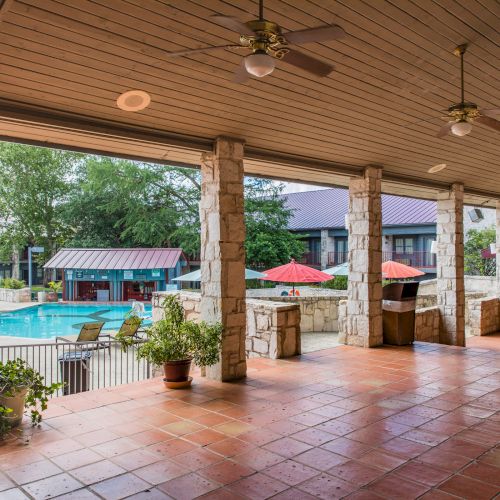 A covered patio with ceiling fans overlooks a swimming pool area, surrounded by trees and red umbrellas, creating a relaxing outdoor space.