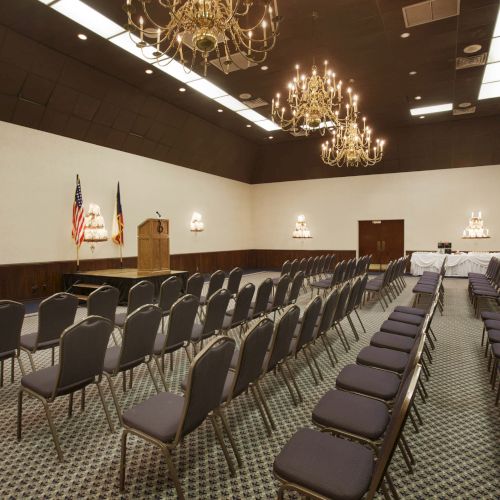 An empty conference room with rows of chairs, a podium, chandeliers, flags, and a carpeted floor.