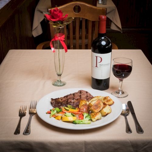A dinner setting with steak, veggies, roasted potatoes, a wine bottle, a glass of wine, and a rose in a vase on a table.