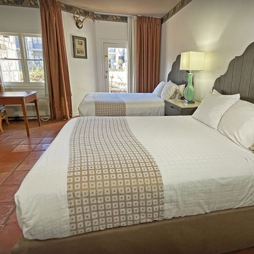 The image shows a hotel room with two beds, a desk, chairs, lamp, and curtains, featuring simple decor and tile flooring.