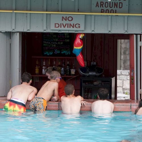 People are in a pool at a swim-up bar with a "No Diving" sign, interacting near drinks. A colorful parrot decoration is visible.