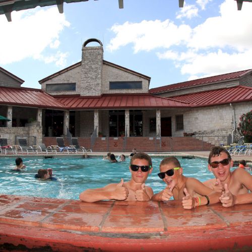 People enjoying a sunny day at a pool, with three individuals at the edge giving thumbs up. A building is in the background, under a blue sky.