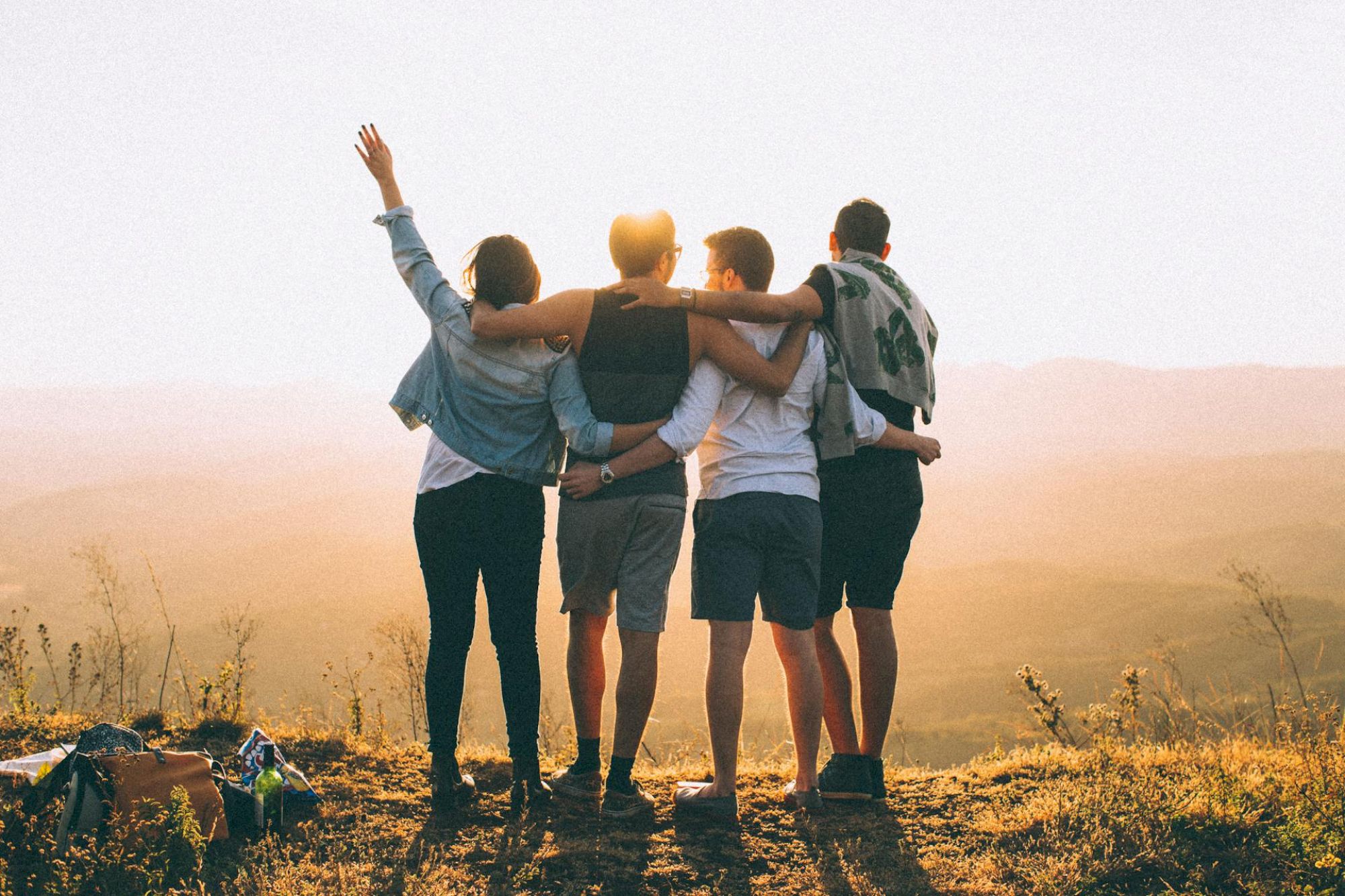 A group of friends stands together on a hill, arms around each other, watching a golden sunset and enjoying the moment.