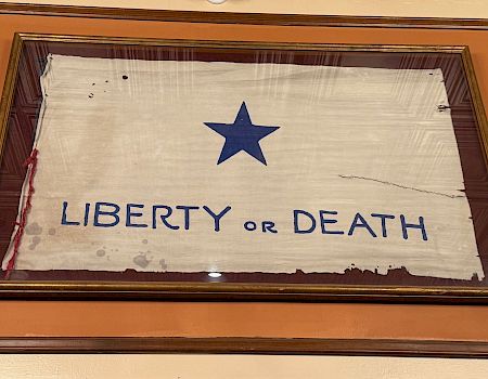 A framed flag with a blue star, the text &ldquo;LIBERTY or DEATH,&rdquo; and a weathered look hangs on an orange wall.