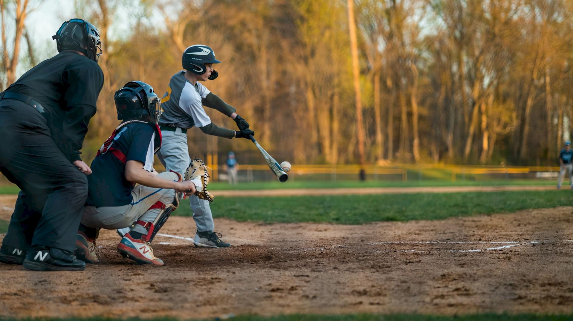 A baseball bat is swung by a batter during a game, with a catcher and umpire nearby and a sunny field in the background.