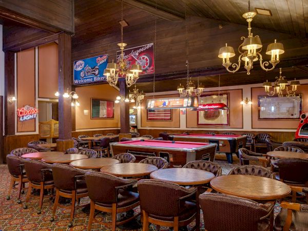 A cozy arcade bar with pool tables, vintage decor, chandeliers, and wall art; warm lighting and many wooden chairs around round tables, inviting.