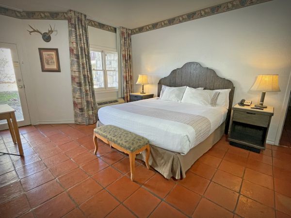 A cozy hotel room with a king bed, matching headboard, two nightstands, lamps, a small bench, terracotta floor, and floral curtains by the window.