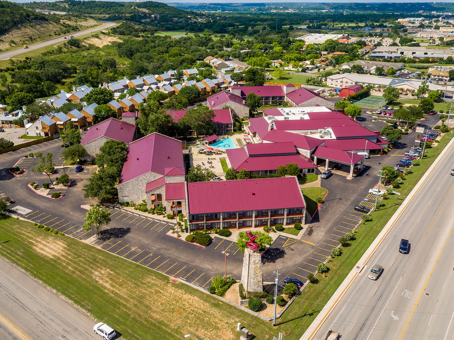 An aerial view of a campus-like complex with red-roofed buildings, parking lots, green lawns, a central monument, and surrounding roads.