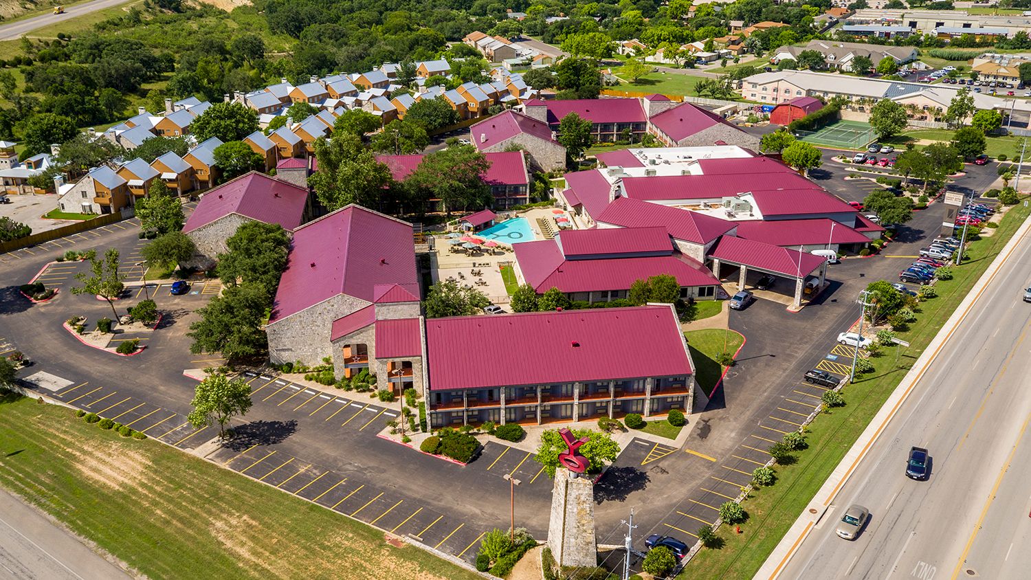 An aerial view of a campus-like complex with red-roofed buildings, parking lots, green lawns, a central monument, and surrounding roads.