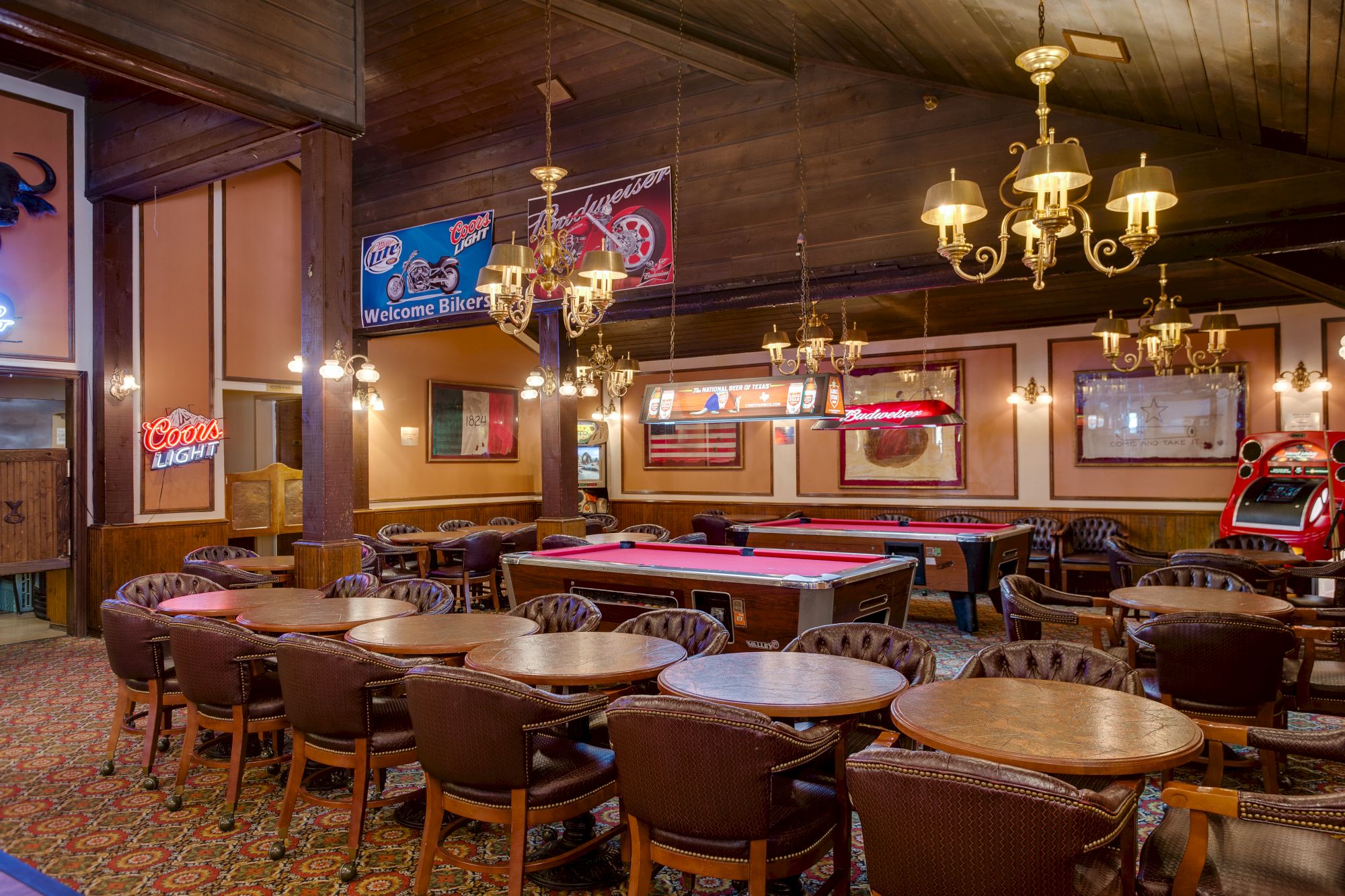 A retro arcade and billiard room with pool tables, vintage chairs, neon signs, chandeliers, and classic game machines.