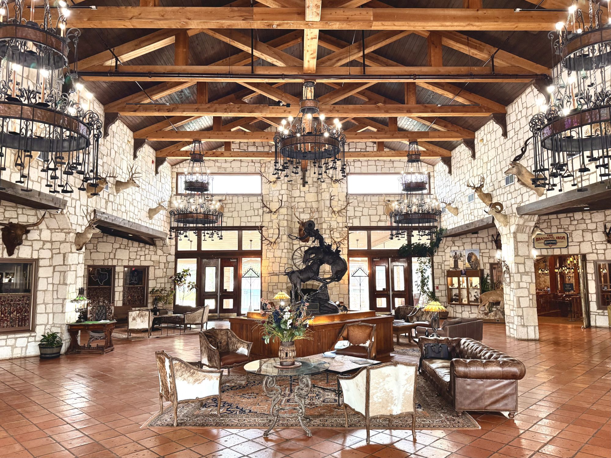 Luxurious stone-walled lobby with high wooden ceilings, chandeliers, leather seating, and a grand statue centerpiece. End.