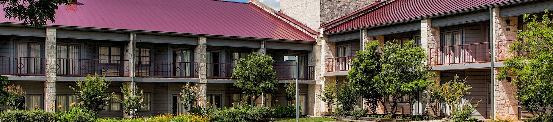 A courtyard with manicured shrubs and walking paths in front of a stone building with red metal roofs under blue skies.