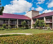 A courtyard with manicured shrubs and walking paths in front of a stone building with red metal roofs under blue skies.