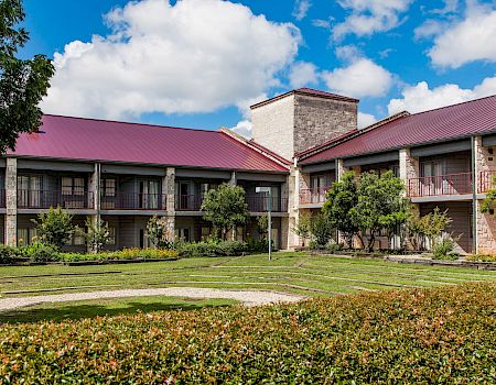 A courtyard with manicured shrubs and walking paths in front of a stone building with red metal roofs under blue skies.