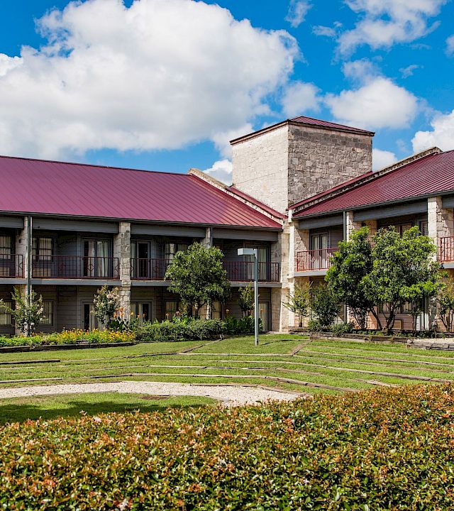 A courtyard with manicured shrubs and walking paths in front of a stone building with red metal roofs under blue skies.