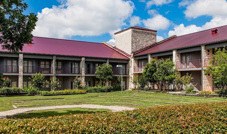 A courtyard with manicured shrubs and walking paths in front of a stone building with red metal roofs under blue skies.