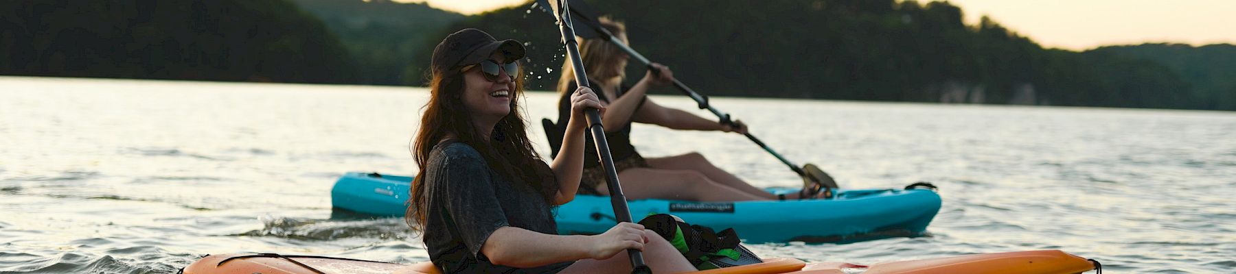 A kayaker paddles an orange canoe at sunset on a calm lake, with another person in the blue kayak behind.