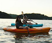 A kayaker paddles an orange canoe at sunset on a calm lake, with another person in the blue kayak behind.