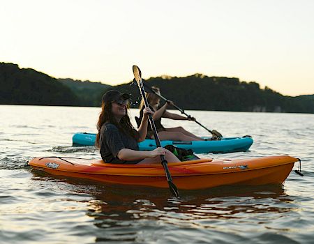 A kayaker paddles an orange canoe at sunset on a calm lake, with another person in the blue kayak behind.
