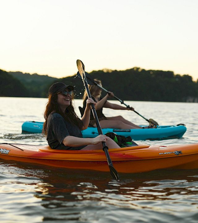 A kayaker paddles an orange canoe at sunset on a calm lake, with another person in the blue kayak behind.