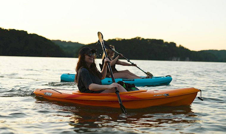 A kayaker paddles an orange canoe at sunset on a calm lake, with another person in the blue kayak behind.