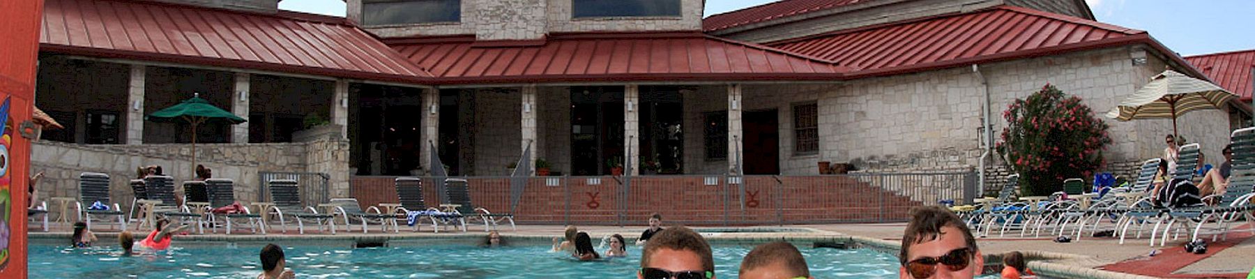 A sunny outdoor pool scene with swimmers lounging by the water and a red-roof building in the background.