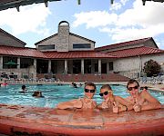 A sunny outdoor pool scene with swimmers lounging by the water and a red-roof building in the background.