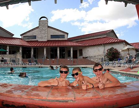 A sunny outdoor pool scene with swimmers lounging by the water and a red-roof building in the background.