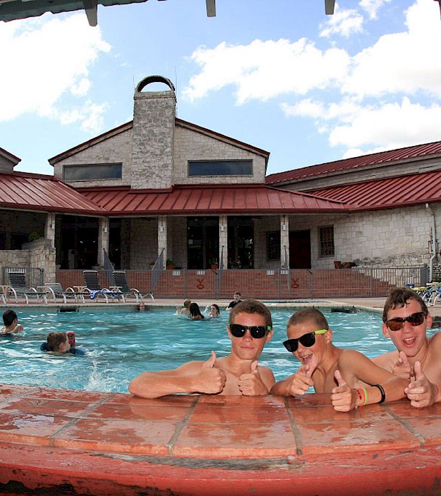 A sunny outdoor pool scene with swimmers lounging by the water and a red-roof building in the background.